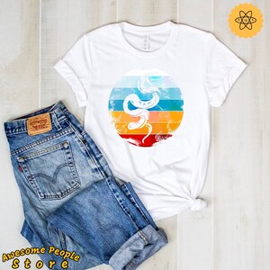 Puede incluir: Camiseta blanca con un gráfico retro de una serpiente con un fondo de puesta de sol colorido.