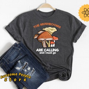 Puede incluir: Una camiseta gris oscuro con un gráfico blanco de setas y el texto "The Mushrooms Are Calling and I Must Go".