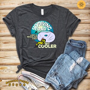 Puede incluir: Una camiseta gris oscuro con un gráfico de un paracaidista con un paracaídas y el texto "Skydiving Dad Like a Regular Dad But Cooler".