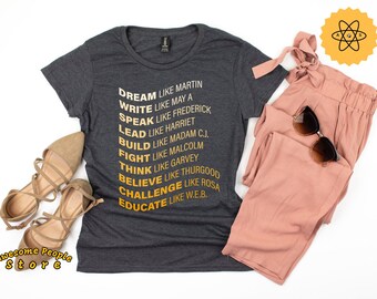 Könnte beinhalten: Ein dunkelgraues T-Shirt mit einem goldenen Aufdruck einer Liste inspirierender Zitate. Der Text lautet "Träume wie Martin, Schreibe wie Maya, Sprich wie Frederick, Führe wie Harriet, Baue wie Madam C.J., Kämpfe wie Malcolm, Denke wie Garvey, Glaube wie Thurgood, Fordere wie Rosa, Bilde wie W.E.B."