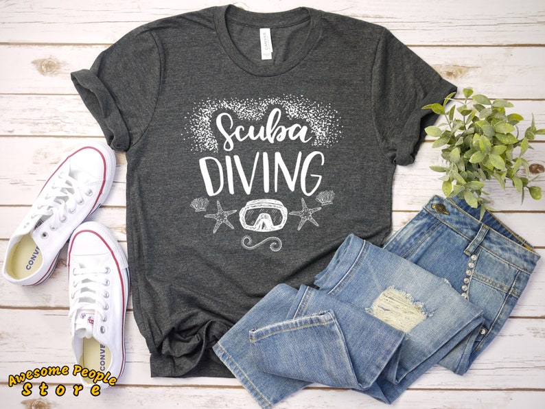 Puede incluir: Una camiseta gris oscuro con el texto "Scuba Diving" en letras blancas. La camiseta tiene un gr&aacute;fico blanco de una m&aacute;scara de buceo y dos estrellas de mar. La camiseta est&aacute; colocada sobre una superficie de madera blanca con un par de zapatillas blancas y unos vaqueros azules.