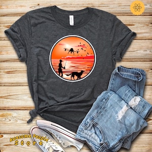 May include: A dark gray t-shirt with a circular design featuring a silhouette of a person fishing with a dog by their side, a drone flying overhead, and a sunset in the background.