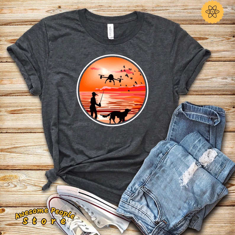May include: A dark gray t-shirt with a circular design featuring a silhouette of a person fishing with a dog by their side, a drone flying overhead, and a sunset in the background.
