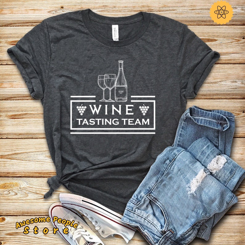 Puede incluir: Una camiseta gris oscuro con un dise&ntilde;o gr&aacute;fico blanco de dos copas de vino y una botella de vino. El gr&aacute;fico tambi&eacute;n incluye el texto "Wine Tasting Team" con un racimo de uvas a cada lado de la palabra "Wine".