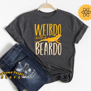 Puede incluir: Camiseta gris oscuro con el texto "WEIRDO with a BEARDO" en amarillo y blanco roto. Un gráfico de lagarto amarillo está encima de la palabra "BEARDO". La camiseta se combina con vaqueros azules. La imagen también incluye una pegatina amarilla con un símbolo científico y el texto "Awesome People Store".