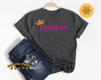 May include: A dark gray t-shirt with a gold crown and the word "Feminist" in pink lettering.