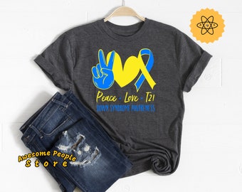 Può includere: Una maglietta grigio scuro con un cuore giallo e un nastro blu. Il testo "Peace + Love + T21 Down Syndrome Awareness" è stampato sulla maglietta.