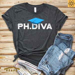Puede incluir: Una camiseta gris oscura con un gráfico de gorro de graduación azul y el texto "PH.DIVA" en blanco con un aspecto desgastado.