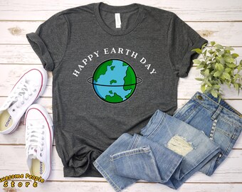 May include: A dark grey t-shirt with the text "Happy Earth Day" and a cartoon image of the Earth in a white circle. The shirt is laid out on a white wooden surface with a pair of white trainers and blue jeans.