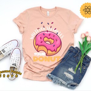 Peut inclure: Un t-shirt rose avec un motif de beigne et le texte "Whatever sprinkles your donut".