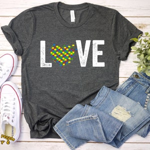 Op de afbeelding: Een donkergrijs T-shirt met het woord "LOVE" in wit gedrukt, met een puzzelhart in het midden van het woord. De puzzelstukjes zijn in de kleuren van het autismebewustzijnlint.