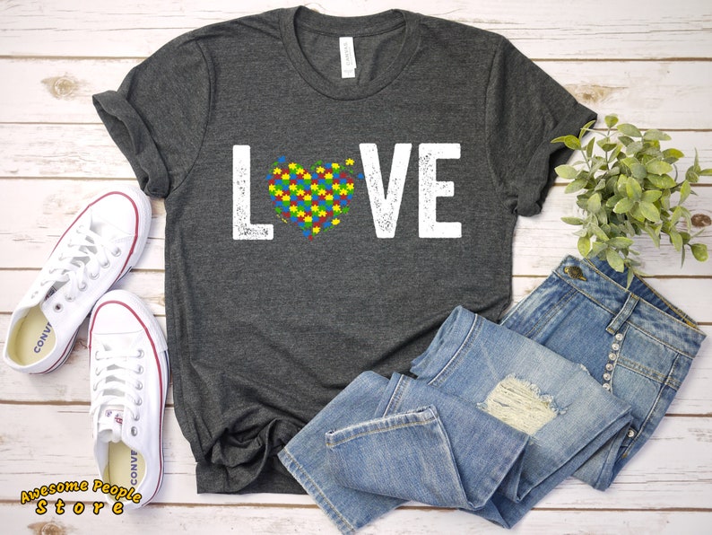 Op de afbeelding: Een donkergrijs T-shirt met het woord "LOVE" in wit gedrukt, met een puzzelhart in het midden van het woord. De puzzelstukjes zijn in de kleuren van het autismebewustzijnlint.