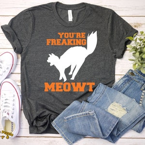 Puede incluir: Una camiseta gris oscuro con el texto "You're Freaking Meowt" en letras naranjas. Una silueta blanca de un gato con la cola levantada est&aacute; por encima del texto. La camiseta est&aacute; colocada sobre una superficie de madera blanca con un par de zapatillas blancas y unos vaqueros azules.