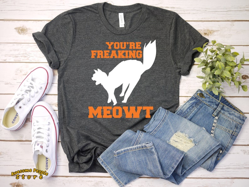 Puede incluir: Una camiseta gris oscuro con el texto "You're Freaking Meowt" en letras naranjas. Una silueta blanca de un gato con la cola levantada est&aacute; por encima del texto. La camiseta est&aacute; colocada sobre una superficie de madera blanca con un par de zapatillas blancas y unos vaqueros azules.