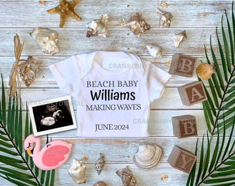 May include: A white onesie with the text "Beach Baby Williams Making Waves June 2024" on it. The onesie is surrounded by seashells, a starfish, a pink flamingo, and wooden blocks that spell out "BABY".