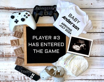Pode incluir: Um quadro de letras preto com o texto "PLAYER #3 HAS ENTERED THE GAME".  Há blocos de madeira que soletram "BABY" e um macacão branco com o texto "BABY Anderson ARRIVING JULY 2024".  Há também uma imagem de ultrassom em preto e branco, uma fralda branca e dois sapatinhos de bebê brancos.
