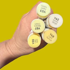 May include: A hand holding a bundle of yellow-toned art markers against a yellow background. The markers are labeled with color names and codes: Y03 Pastel Yellow, Y07 Banana Cream, Y26 Light Gold, Y48 Light Apricot, and Y62 Sandy Beige.