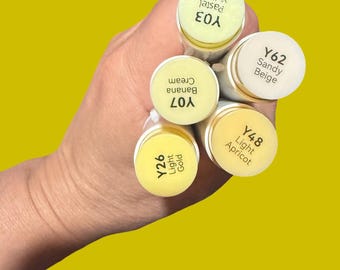Puede incluir: Primer plano de una mano sosteniendo un conjunto de rotuladores de arte en tonos amarillos sobre un fondo amarillo. Los rotuladores están etiquetados con nombres y códigos de color: Y03 Pastel Yellow, Y07 Banana Cream, Y26 Light Gold, Y48 Light Apricot y Y62 Sandy Beige.