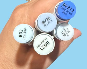 Puede incluir: Una mano sosteniendo una colección de rotuladores de arte de colores. Los rotuladores tienen cuerpos blancos y tapas de colores con etiquetas de texto como "Frost Blue", "Sky Blue" y "Deep Royal Blue". El fondo es azul claro liso.