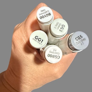 May include: A hand holding five art markers with white barrels and gray caps. The caps are labeled with color names and codes, including "GG1 Green Grey 1", "GG3 Green Grey 3", and "CG5 Cool Grey #5". The background is a solid gray.