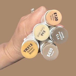 May include: A hand holding several art markers with various shades of brown and beige. The markers are labeled with color codes and names, including "E120 Reddish Brass," "E46 Coral Sand," and "YR313 Potato Brown." The background is a solid brown.