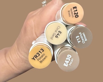 Puede incluir: Una mano sosteniendo varios rotuladores artísticos en tonos marrones y beige. Los rotuladores están etiquetados con códigos de color y nombres, incluyendo "E120 Reddish Brass", "E46 Coral Sand" y "YR313 Potato Brown". El fondo es marrón liso.