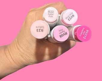 Puede incluir: Una mano sostiene cinco rotuladores artísticos de color rosa sobre un fondo rosa. Los rotuladores tienen barriles blancos y tapas rosas con etiquetas: R22 Ballet Pink, RV23 Pink Wisp, RV212 Rich Raspberry, RV08 Pastel Pink y R23 Sakura.