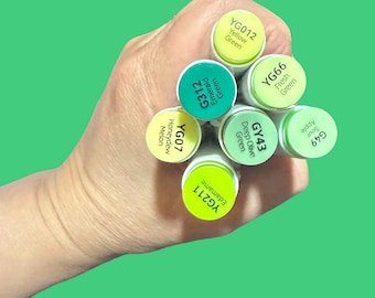 Puede incluir: Una colección de rotuladores de arte en tonos verdes, incluyendo tonos como Verde Amarillo, Verde Esmeralda y Verde Fresco. Cada rotulador tiene una tapa con el nombre y código del color. Los rotuladores están sobre un fondo verde.