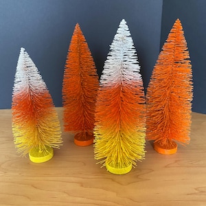 May include: Four decorative bottle brush trees in the shape of candy corn. The trees are orange, yellow, and white, with a round base. The trees are arranged on a wooden surface with a dark blue background.