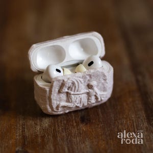 Puede incluir: Una funda blanca para AirPods con una cubierta beige claro texturizada con un diseño de escultura en relieve. La funda está abierta, revelando auriculares blancos en el interior. El fondo es una superficie de madera borrosa. El logo de Alexa Roda es visible.