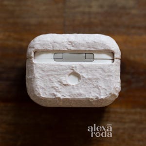 K&ouml;nnte beinhalten: Eine hellbeige strukturierte H&uuml;lle f&uuml;r kabellose Ohrh&ouml;rer. Die H&uuml;lle hat eine rechteckige Form mit abgerundeten Kanten und einer kleinen kreisf&ouml;rmigen &Ouml;ffnung an der Unterseite. Die Oberseite der H&uuml;lle hat einen silberfarbenen Abschnitt. Der Markenname "alexa roda" ist unten rechts sichtbar.