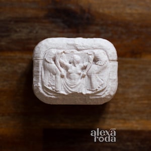 K&ouml;nnte beinhalten: Kleine, cremefarbene, rechteckige Box mit einem geschnitzten Relief, das drei Figuren darstellt. Die zentrale Figur wird von zwei anderen flankiert, alle in klassische Gew&auml;nder geh&uuml;llt. Die Box hat abgerundete Kanten und eine strukturierte Oberfl&auml;che. Das Logo "alexa roda" ist sichtbar.