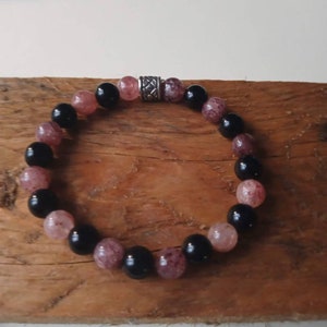 May include: A black and pink beaded bracelet with a silver charm. The bracelet is made of round beads and has a simple design.