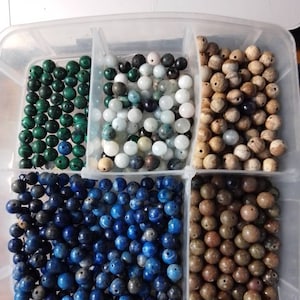 May include: A clear plastic container with six compartments filled with various colored beads. The beads are in shades of green, blue, brown, and white.