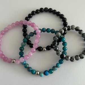 May include: Four beaded bracelets in pink, black, teal, and grey. Each bracelet is made of small, round beads strung together. The bracelets are arranged in an overlapping pattern, with a silver bead accent.