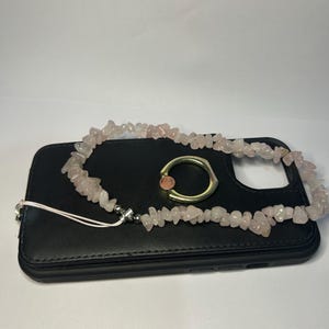 Puede incluir: Una funda de teléfono negra con una correa para teléfono de cuentas de cuarzo rosa y un soporte para anillo de teléfono dorado. La correa está hecha de pequeñas piedras de cuarzo rosa claro. El soporte del anillo tiene una pequeña piedra rosa en el centro.