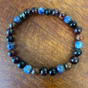 Può includere: Un braccialetto di perline con perline di pietre preziose nei toni del blu, marrone e nero. Le perline rotonde sono infilate insieme per formare un braccialetto circolare. Le perline hanno una finitura lucida. Il braccialetto è adatto all'uso quotidiano.