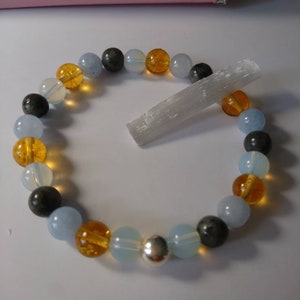 May include: A bracelet made of blue, gray, and orange beads with a silver charm. The bracelet is lying on a white surface next to a white crystal.