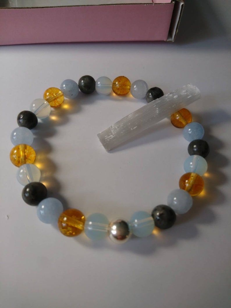 May include: A bracelet made of blue, gray, and orange beads with a silver charm. The bracelet is lying on a white surface next to a white crystal.