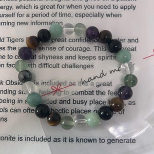 May include: A bracelet made with various gemstones, including amethyst, tiger's eye, clear quartz, green aventurine, black obsidian, and fluorite. The bracelet is on a white background with text that reads "hand made".