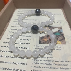 May include: A white and black beaded bracelet with two hematite balls. The bracelet is in a box with a card that reads "Selenite" and lists the benefits of the stone. The benefits include: removes negative energies, relieves anxiety, brings deep peace, unlocks memory of past and future, enhances psychic development, angelic communication & higher guidance, encourages self-awareness.