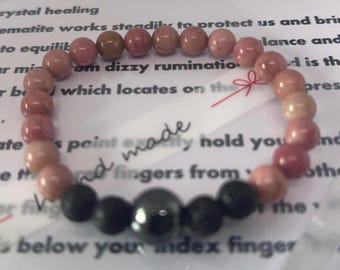 Könnte beinhalten: Ein rosa-schwarzes Armband mit einem silbernen Anhänger. Das Armband ist aus natürlichen Steinen gefertigt und hat ein handgeschriebenes Etikett mit der Aufschrift "handgefertigt".