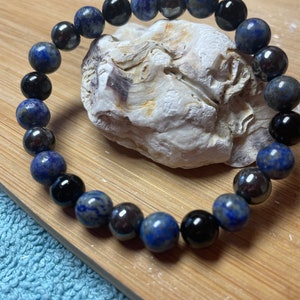 Puede incluir: Un brazalete hecho de cuentas negras y azules. Las cuentas son redondas y tienen una superficie lisa. El brazalete está sobre una superficie de madera.