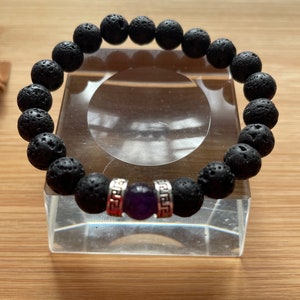 May include: Black lava rock beaded bracelet with a purple amethyst gemstone and silver accents. The bracelet has a Greek key design on the silver accents.