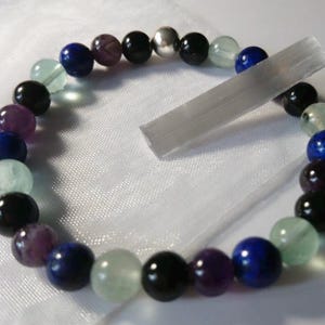 Puede incluir: Un brazalete hecho de cuentas negras, amatistas, verdes y azules con un dije de plata. El brazalete está sobre un fondo blanco con un cristal transparente al lado.
