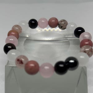 May include: A beaded bracelet featuring a variety of gemstone beads in shades of pink, white, black, and brown. The bracelet is displayed on a clear, angled surface, showcasing the arrangement of the round beads.