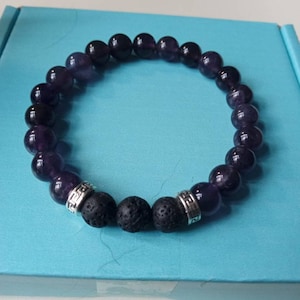 Puede incluir: Una pulsera de cuentas con cuentas de amatista morada y piedra de lava negra. Las cuentas de amatista son de color púrpura intenso y las cuentas de lava son negras. También se incluyen espaciadores metálicos plateados. La pulsera está sobre una superficie azul claro.