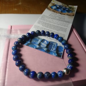 May include: A bracelet featuring dark blue lapis lazuli beads and a silver bead. The bracelet is presented on a pink box, accompanied by a card with the text "LAPIS LAZULI SELF-EXPRESSION WISDOM TRUTH".