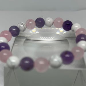 Könnte beinhalten: Ein Stretch-Armband mit runden Perlen in Lila, Rosa und Weiß. Das Armband besteht aus Amethyst-, Rosenquarz- und weißem Howlith. Die Perlen sind auf einem elastischen Band aufgereiht.