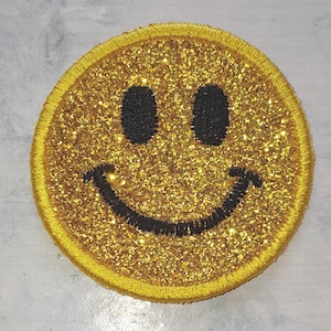 Può includere: Toppa con faccina sorridente glitterata dorata con bordo giallo. Gli occhi sono ovali neri e la bocca è una linea nera curva. La toppa è rotonda e ha una superficie lucida e strutturata.